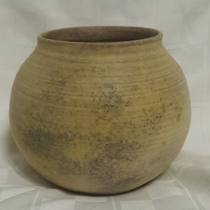 May include: A round, handmade ceramic vase with a wide opening. The vase has a light yellow color with darker, speckled markings. The exterior features horizontal, textured lines. The vase is suitable for home decor.
