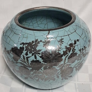 May include: A round, turquoise ceramic vase with a silver rim and floral design. The vase has a crackle glaze effect, and the flowers are in a dark silver color. The vase is a decorative item.