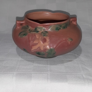 May include: A matte terracotta-colored ceramic bowl featuring two small side handles and a raised relief design of light pink flowers and dark green leaves encircling the exterior. The bowl has a wide, open mouth and a rounded, bulbous body shape.