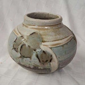 May include: A handcrafted ceramic vase with a round body and a wide opening. The vase features a textured surface with a mix of colors, including light blue, beige, and brown. Decorative leaf-like patterns are carved into the surface.