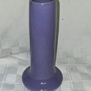 May include: A tall, cylindrical purple ceramic vase with a smooth, glossy finish. The vase features a wide, circular base that tapers upward into a slender, straight body with a simple, open rim at the top, showcasing a solid, uniform color.
