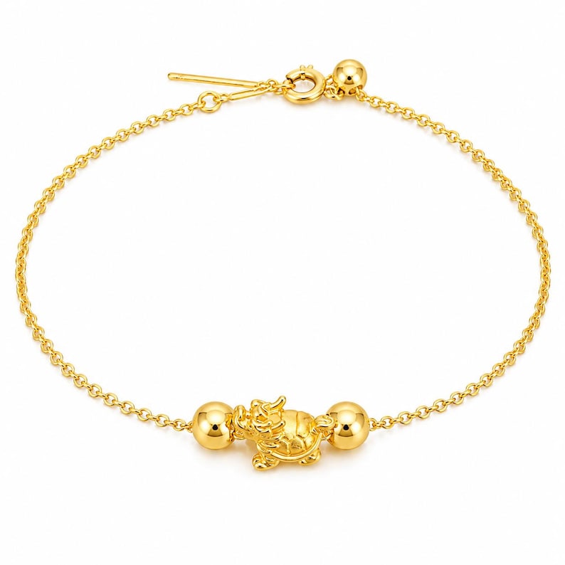 May include: A gold chain bracelet featuring a central charm shaped like a mythical dragon-turtle creature. The charm is flanked by two polished gold spheres on either side. The bracelet is secured with a circular spring ring clasp and a small bead.