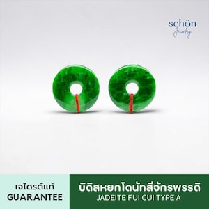 Peut inclure: Deux pendentifs circulaires en jadéite verte en forme de beignet avec une finition lisse et polie. Chaque pièce présente un petit trou central et un fin cordon rouge attaché au bord inférieur. Le texte "schön Jewelry" est visible en haut.