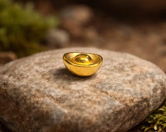 May include: A small, gold-colored ingot, resembling a traditional Chinese Yuan Bao, rests on a textured, gray stone. The ingot has a rounded shape with a central, raised area. The background features blurred green foliage and a natural, earthy setting.