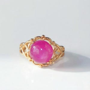 May include: A gold-colored ring featuring a large, round, vibrant pink gemstone. The gemstone is set in an ornate, filigree-style setting with small accent stones. The ring is displayed on a white surface.