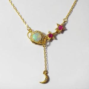 May include: A gold necklace with a celestial theme. It features a central opal-like stone encircled by small clear stones, a chain leading to a crescent moon pendant, and star-shaped accents with red gemstones. The necklace is a delicate piece.