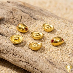 May include: Six gold-colored, ingot-shaped beads with a textured surface are arranged on a piece of driftwood and sand. Two beads have red holes. The beads are likely for jewelry making or crafting.