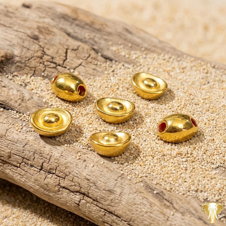 May include: Six gold-colored, ingot-shaped beads with a textured surface are arranged on a piece of driftwood and sand. Two beads have red holes. The beads are likely for jewelry making or crafting.