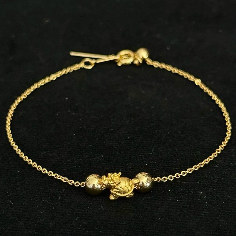 May include: A delicate gold chain bracelet featuring a central charm shaped like a small turtle, flanked by two smooth, polished gold spherical beads. The bracelet is secured with a gold spring ring clasp and a short extender chain at the end.