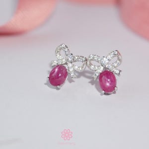 May include: A pair of silver bow-shaped earrings with oval, deep pink gemstones. The bows are encrusted with small, clear gemstones. The earrings are set against a soft, white background with a blurred pink ribbon in the background.