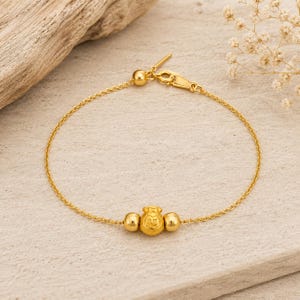 May include: A gold chain bracelet featuring a central charm shaped like a small money bag with the Chinese character for good fortune engraved on it, flanked by two small gold spheres and secured with a gold lobster claw clasp.
