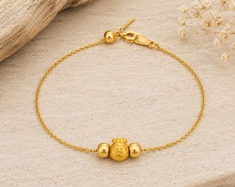 May include: A gold chain bracelet featuring a central charm shaped like a small money bag with the Chinese character for good fortune engraved on it, flanked by two small gold spheres and secured with a gold lobster claw clasp.
