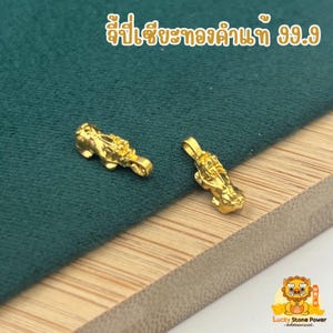 Peut inclure: Deux pendentifs dorés, chacun en forme de créature mythique, sont présentés sur une surface verte. Les pendentifs présentent des détails complexes et une finition brillante. Du texte thaïlandais est visible en haut de l'image.