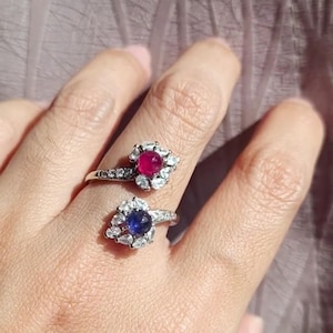 Peut inclure: Deux bagues en argent, chacune ornée d'une pierre précieuse centrale entourée de petites pierres. L'une a une pierre rouge, l'autre une bleue. Les bagues sont portées sur un doigt, mettant en valeur le design des bijoux.