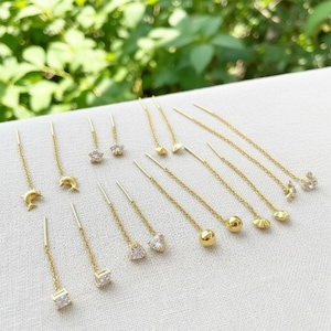 Può includere: Una collezione di orecchini a filo color oro con vari ciondoli, tra cui delfini, stelle, sfere e cristalli trasparenti. Ogni orecchino è composto da una sottile catenina dorata progettata per essere infilata attraverso il lobo dell'orecchio.