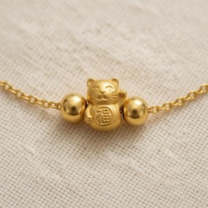 May include: A gold-toned chain bracelet featuring a central charm shaped like a beckoning cat with a raised paw. The cat charm is flanked by two smooth, spherical gold-toned beads. The cat figure has a small square symbol engraved on its chest.