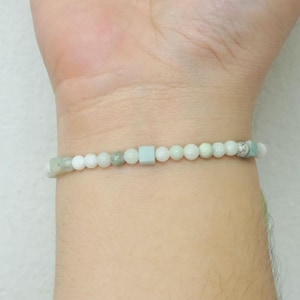 Könnte beinhalten: Ein zartes Armband aus kleinen, runden und quadratischen Perlen in Weiß-, Grün- und Hellblautönen. Das Armband wird am Handgelenk getragen und zeigt seine subtile Farbpalette und sein minimalistisches Design. Die Perlen haben einen Durchmesser von etwa 0,5 cm.