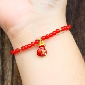 Op de afbeelding: Rode kralenarmband met een gouden bedel in de vorm van een kleine tas. De armband zit om een pols. De bedel heeft gouden accenten en witte details. De armband is gemaakt van kleine, ronde rode kralen.