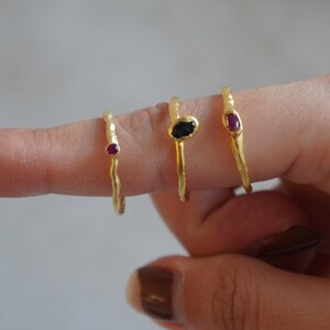 May include: Three gold rings with textured bands, each featuring a different gemstone. One ring has a small, oval-shaped ruby, another has a dark, irregular-shaped stone, and the third has a small, oval-shaped ruby.
