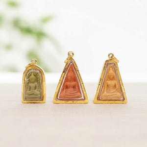 Peut inclure: Trois pendentifs représentant des bouddhas assis dans des cadres dorés. Le pendentif de gauche est arrondi avec une figurine vert sauge, tandis que les deux de droite sont triangulaires avec des figurines couleur terre cuite et pêche.