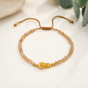 May include: A bracelet featuring small, translucent, light brown rutilated quartz beads strung on a brown cord. The center displays a detailed gold-colored charm shaped like a mythical creature, flanked by two small gold-colored beads. It has an adjustable sliding knot closure.
