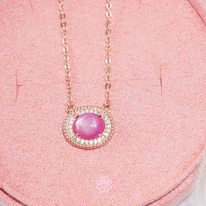 May include: A rose gold necklace featuring a pink gemstone pendant. The pendant is oval-shaped, surrounded by a halo of small, clear stones. The necklace chain is delicate and complements the pendant's design. The background is a soft pink.