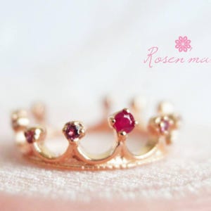 May include: A gold-colored crown ring with several red gemstones. The ring has a delicate design with small, rounded points and a central, larger gemstone. The ring is set against a soft, pink background, with the text "Rosen ma" in pink.