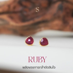 Peut inclure: Boucles d'oreilles à clous dorées avec des pierres précieuses rubis rouge foncé triangulaires. Le mot "RUBY" est affiché en rose en dessous. Le fond est blanc avec un élément en bois flou.