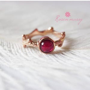 Peut inclure: Une bague en or rose avec une pierre précieuse rouge foncé et ronde. La monture de la bague a un design texturé en forme de brindille. Le texte "Rosen marry" est écrit en rose. La bague est posée sur un fond blanc et vaporeux.