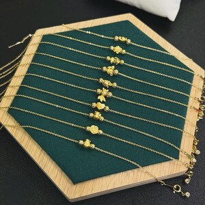 May include: A collection of ten delicate gold chain bracelets, each featuring a unique central charm including shapes like a heart, a flower, and various small figures, all arranged in parallel lines on a dark green hexagonal display base.