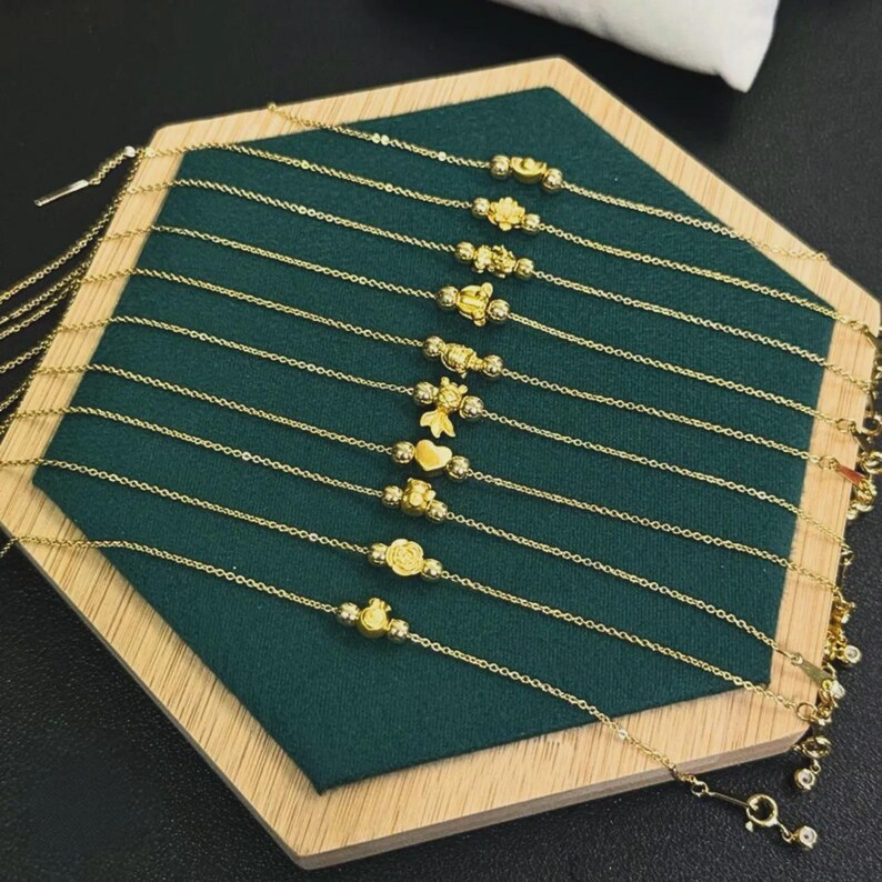 May include: A collection of ten delicate gold chain bracelets, each featuring a unique central charm including shapes like a heart, a flower, and various small figures, all arranged in parallel lines on a dark green hexagonal display base.