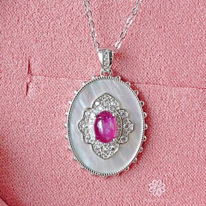 Può includere: Una collana d'argento con un ciondolo con una pietra preziosa rosa circondata da piccole pietre trasparenti. Il ciondolo è incastonato in una forma ovale bianca con un bordo decorativo. La collana è esposta su una superficie rosa.
