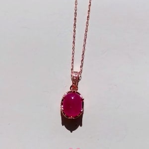 May include: A rose gold necklace with a magenta oval gemstone pendant. The pendant is set in a decorative bezel and hangs from a delicate chain. The necklace is displayed against a white background. The chain is made of rose gold.