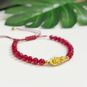 May include: A red beaded bracelet with a gold charm. The bracelet features small, faceted red beads strung together. A gold charm with intricate details is centered on the bracelet. The bracelet is on a white surface with green leaves in the background.