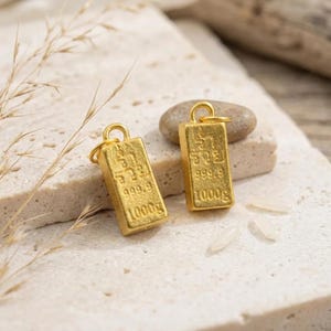 May include: Two rectangular gold-colored metal charms shaped like miniature bullion bars. Each charm features a small circular jump ring at the top and is embossed with the text 999.9 and 1000g, along with additional characters.