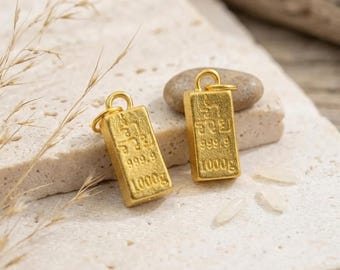 May include: Two rectangular gold-colored metal charms shaped like miniature bullion bars. Each charm features a small circular jump ring at the top and is embossed with the text 999.9 and 1000g, along with additional characters.