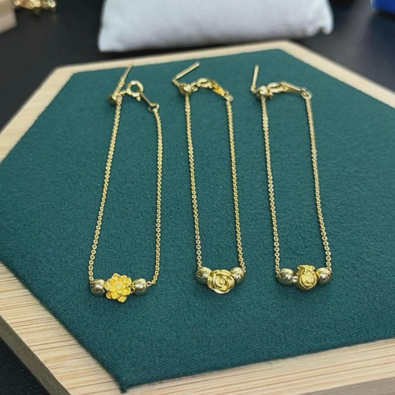 May include: Three gold-colored bracelets, each featuring a delicate chain and a central charm. The left charm is a flower, the middle is a rose, and the right is a small bag. Each bracelet includes two small gold-colored beads flanking the central charm.