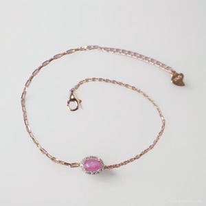 Könnte beinhalten: Roségoldenes Armband mit einem ovalen, rosa Edelstein, umgeben von kleinen, klaren Steinen. Die feine Kette hat einen kleinen Verschluss und ein herzförmiges Etikett. Das Armband wird vor weißem Hintergrund gezeigt.