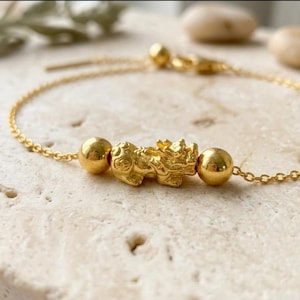 Peut inclure: Un bracelet à chaîne dorée orné d'une breloque centrale en forme de créature mythique, encadrée par deux perles dorées lisses et rondes. Le bracelet présente une fine chaîne à maillons et un petit fermoir doré visible en haut du cadre.