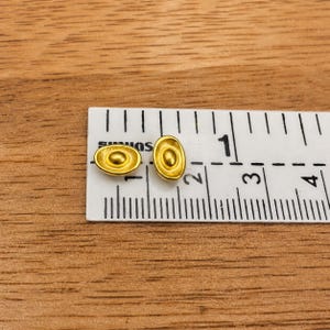 May include: Two small, gold-colored oval-shaped earrings with a central circular design. The earrings are next to a ruler, which measures them at approximately 0.25 inches in length. The earrings are on a wooden surface.