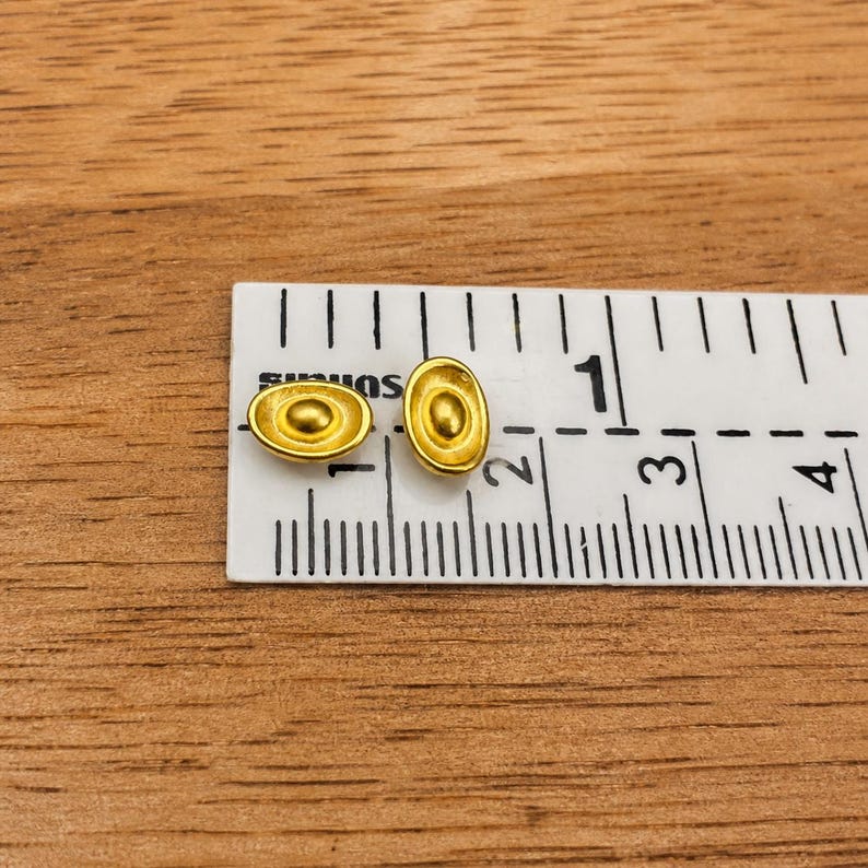 May include: Two small, gold-colored oval-shaped earrings with a central circular design. The earrings are next to a ruler, which measures them at approximately 0.25 inches in length. The earrings are on a wooden surface.
