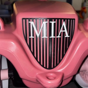 May include: A pink toy car featuring the name "MIA" in white on the front grille. The car has large, cartoon eyes and black wheels. The design is playful and suitable for children. The car's measurements are in centimeters.