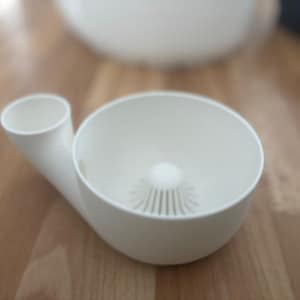 Könnte beinhalten: Ein weißer Zitruspresser mit einer Schüssel, einem Sieb und einem Ausguss. Der Entsafter besteht aus Kunststoff und steht auf einer Holzoberfläche. Das Design umfasst einen kleinen, zylindrischen Ausguss, der sich von der Seite der Schüssel erstreckt.