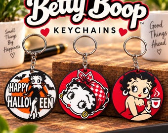 Puede incluir: Tres llaveros circulares de Betty Boop con anillas y cadenas de metal. El llavero de la izquierda muestra a Betty Boop con un vestido negro y el texto Happy Halloween. El centro muestra su rostro con un pañuelo rojo. A la derecha, sostiene una taza.