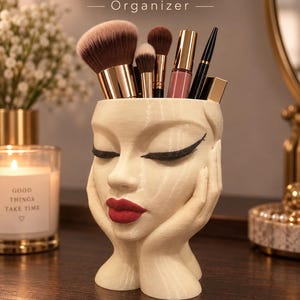 Peut inclure: Un organisateur imprimé en 3D de couleur crème en forme de tête humaine avec les mains posées sur les joues. Le visage présente un trait d'eye-liner noir et des lèvres rouges. Il contient des pinceaux de maquillage, un rouge à lèvres et un crayon.