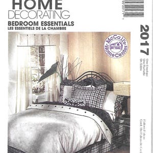 May include: McCall's Home Decorating Bedroom Essentials sewing pattern envelope 2017 featuring a bed with a plaid bed skirt, plaid pillow shams, a solid light-colored duvet with a dark trim, and a matching plaid throw pillow in a bedroom setting.