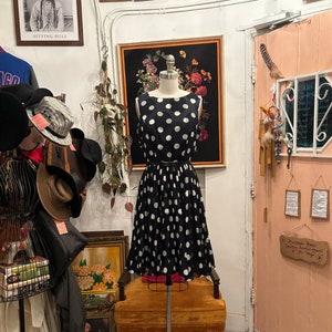 May include: A black and white polka dot dress on a mannequin in a shop. The dress has a fitted bodice and a full skirt. The mannequin is standing on a rolling stand in front of a yellow chair.