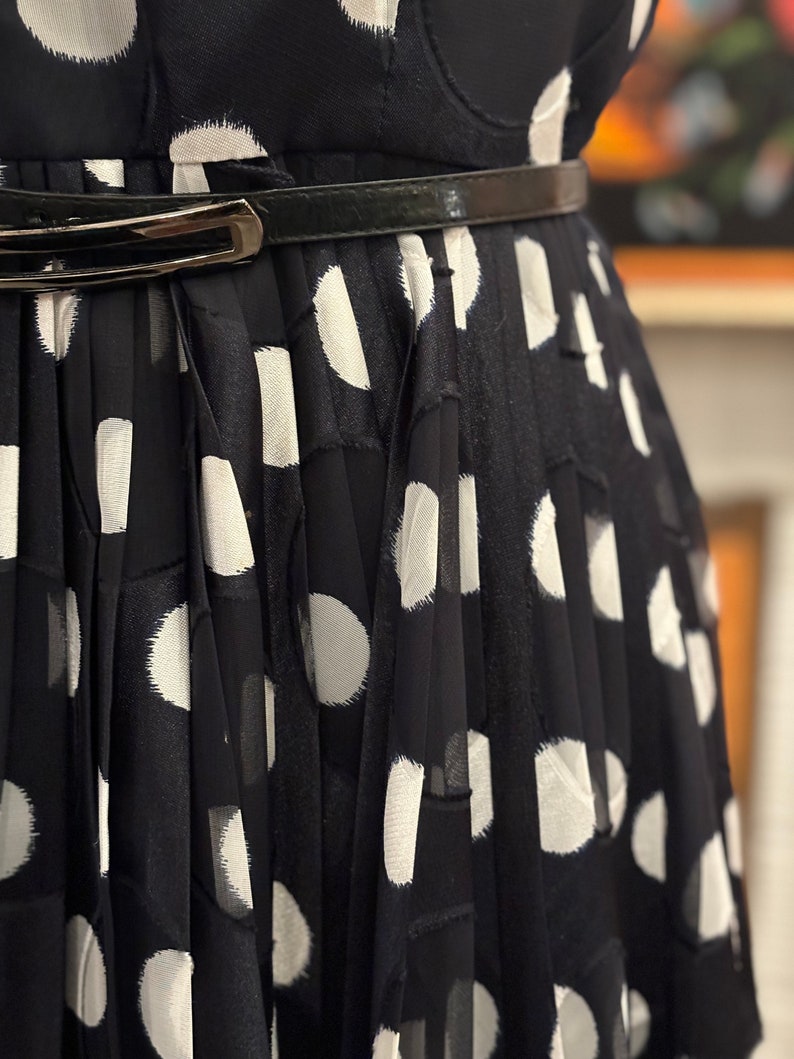 May include: A black and white polka dot dress with a black belt. The dress is pleated and has a loose fit.