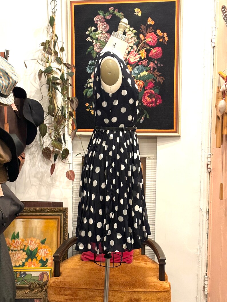 May include: A black and white polka dot dress with a black belt. The dress is on a mannequin and is in a store setting.