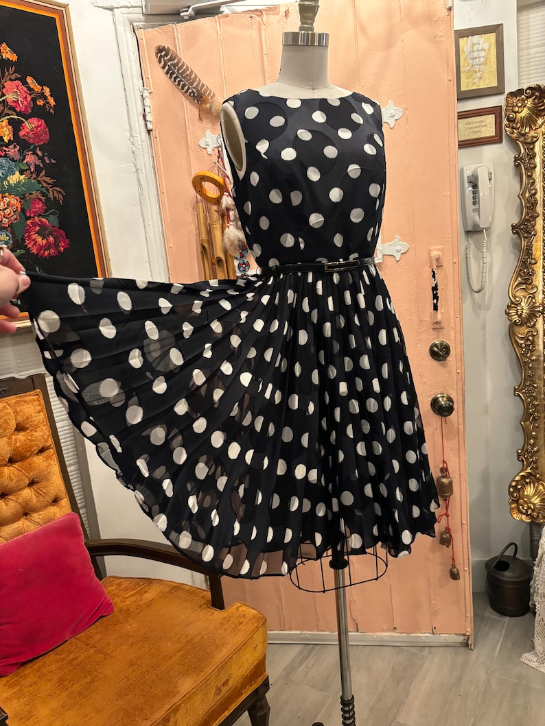 May include: A black and white polka dot dress with a fitted bodice and a full skirt. The dress has a black belt and is sleeveless.